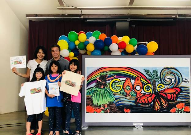 A family of five stands beside a large framed artwork featuring a colorful rainbow, butterfly, and the Google logo. Balloon decorations and award items surround the group as they pose on a stage during a student art recognition event.