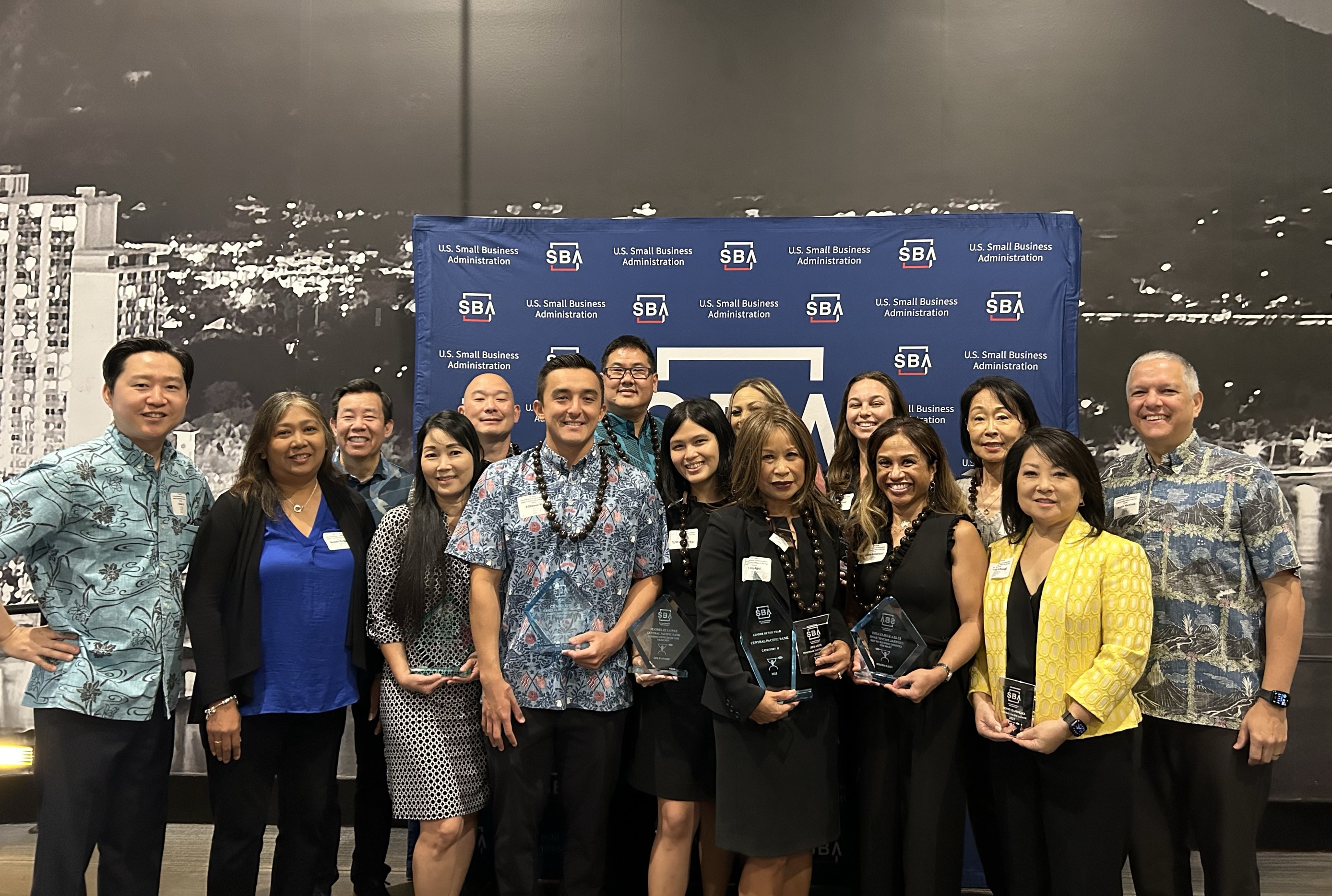 A group of professionals poses in front of a U.S. Small Business Administration (SBA) backdrop, with several individuals holding award plaques. The group photo appears to be taken during an SBA recognition event celebrating business and community achievements.